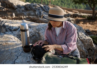 A woman spends time outdoors, sitting on rocky terrain with a backpack and thermos beside her. She is dressed casually in a hat and plaid shirt, preparing for a hike or adventure. - Powered by Shutterstock