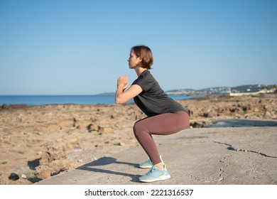 Woman In Sportswear Doing Squat Exercise At Seaside. Female Practicing Workout During Sunny Day