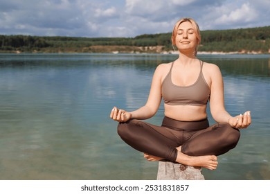 Woman in sportswear is doing yoga while sitting in the lotus position on a wooden beam at the lake - Powered by Shutterstock