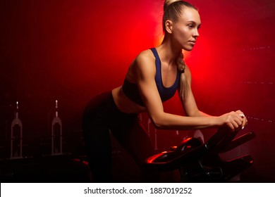 Woman In Sportswear Exercising On Bicycle At Modern Gym, Workout Woman Training On Smart Stationary Bike Indoors In Red Neon Lighted Smoky Space