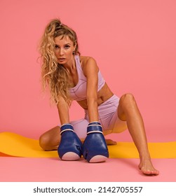 Woman In Sportswear Posing With Boxing Gloves Sitting On Mat On Pink Background