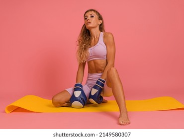 Woman In Sportswear Posing With Boxing Gloves Sitting On Mat On Pink Background