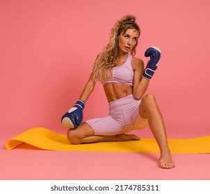 Woman In Sportswear Posing With Boxing Gloves Sitting On Mat On Pink Background