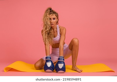 Woman In Sportswear Posing With Boxing Gloves Sitting On Mat On Pink Background