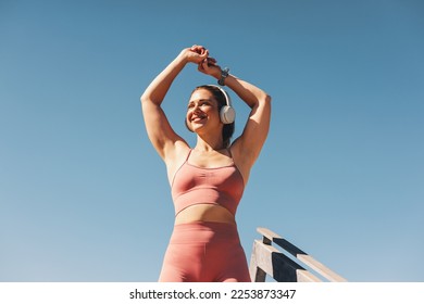 Woman in sportswear stretching her arms outdoors. Happy sportswoman doing a warm up exercise with headphones on. Female athlete working out against the sky. - Powered by Shutterstock