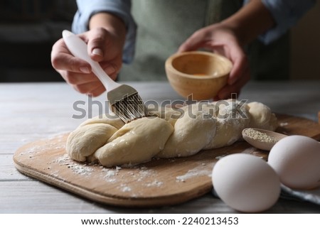 Woman spreading egg yolk onto raw braided bread at white wooden table, closeup. Traditional Shabbat challah