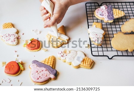 Woman squeezing icing cover on cookies from a pastry bag. Decorating sugar cookies in lolly ice cream shape with sugar glaze. Summer concept