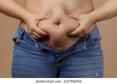 Woman squeezing showing fat sagging tummy in blue unzipped jeans on beige background. Sudden weight gain. Visceral fat. Body positive. Tight little clothes. Need for wardrobe change.