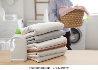 Woman with stack of clean bed sheets and bottle of detergent on wooden table in bathroom