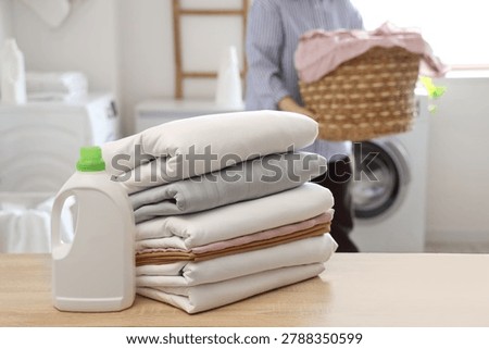 Woman with stack of clean bed sheets and bottle of detergent on wooden table in bathroom