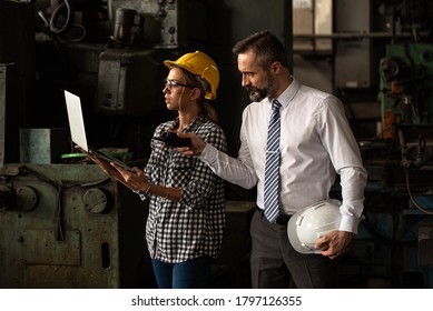 Woman Staff Factory Worker With Hardhat Or Helmet And Engineering Manager In Suit Using Computer And Laptop Discussing Manufacturing Plan Working Inside Industrial Plant