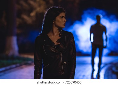 The Woman Stand On A Dark Alley On A Background Of The Man In Fume. Night Time