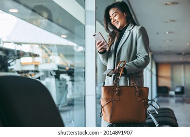 Woman Standing At Airport Lounge Looking At Her Mobile Phone And Smiling. Female Business Traveler At Airport Waiting For The Flight.