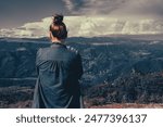 A woman, standing alone with her back to the camera on a hilltop, gazing at picturesque mountains under a dramatic cloudy sky.