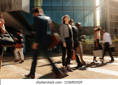 Woman Standing Amidst A Busy Office Going Crowd Hooked To Their Mobile Phones. Businesswoman Holding Her Hand Bag Standing Still On A Busy Street With People Walking Past Her Using Mobile Phones.