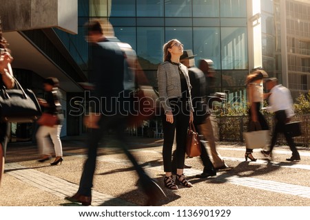 Woman standing amidst a busy office going crowd hooked to their mobile phones. Businesswoman holding her hand bag standing still on a busy street with people walking past her using mobile phones.