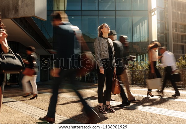 Woman standing amidst a busy office going crowd\
hooked to their mobile phones. Businesswoman holding her hand bag\
standing still on a busy street with people walking past her using\
mobile phones.