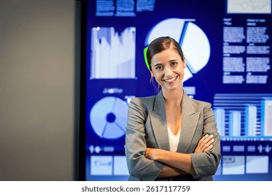 Woman standing arms crossed and smiling in office, digital screen showing pie charts and bar graphs. Professional, executive, corporate, data visualization, analytics, presentation, leadership