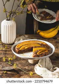 Woman Standing In Background Eating Banana Upside Down Cake With Banana Bread Sliced On Rustic Wooden Table On A White Plate 