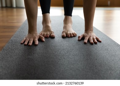 Woman standing barefoot on black rubber mat performs Standing Forward Bend, doing Uttanasana exercise, cropped body part view, palms and feet close up image. Yoga asana, training, sport class concept