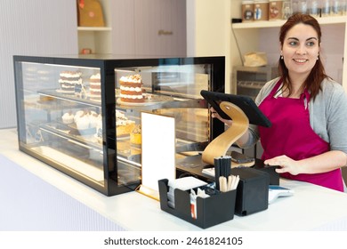 A woman is standing behind a counter in a bakery, smiling and holding a tablet. The display case behind her is filled with cakes and pastries, including a cake with a frosting that looks like a cake - Powered by Shutterstock