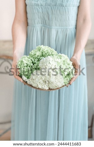 A woman standing in a blue dress, holding a bowl filled with colorful flowers.