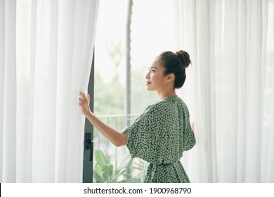 Woman Standing By Living Room Window And Opening Curtains