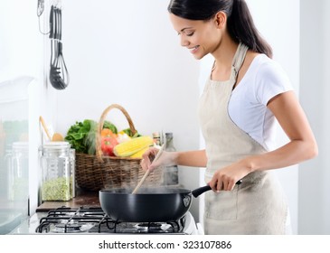 Woman standing by the stove in the kitchen, cooking and smelling the nice aromas from her meal in a pot - Shutterstock ID 323107886