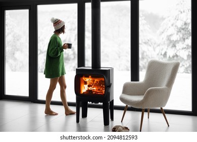 Woman Standing With Cup By The Fireplace Near The Window At Modern House On Nature During Winter Time. Concept Of Winter Mood And Comfort At Home. Idea Of Recreation In Cabins On Mountains