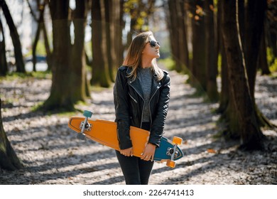 Woman standing at forest alley and carrying longboard at summer time. Skater in casual wear hold longboard in hands 