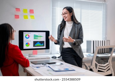 A woman is standing in front of a computer monitor and talking to another woman. The woman on the left is wearing a red shirt and the woman on the right is wearing a white shirt