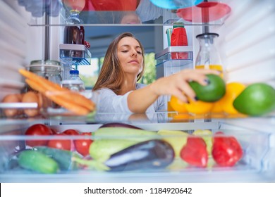 Woman Standing In Front Of Opened Fridge And Taking Avocado. Fridge Full Of Groceries.