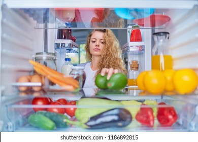 Woman Standing In Front Of Opened Fridge And Taking Avocado. Fridge Full Of Groceries.