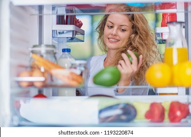 Woman Standing In Front Of Opened Fridge And Taking Avocado. Fridge Full Of Groceries. Picture Taken From The Iside Of Fridge.