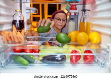 Woman Standing In Front Of Opened Fridge And Taking Avocado. Fridge Full Of Groceries. Picture Taken From The Iside Of Fridge.