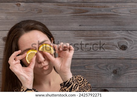 A woman standing in front of a wall made of old wooden boards and holding lemons half-slices over her eyes.
