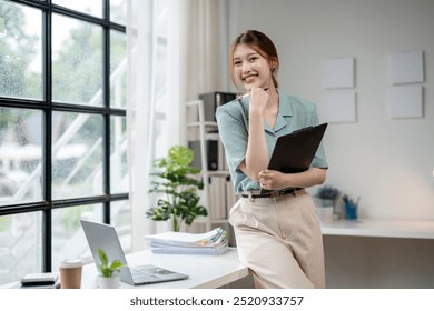 A woman is standing in front of a window with a laptop and a clipboard. She is smiling and she is in a professional setting - Powered by Shutterstock