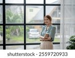 A woman is standing in front of a window with rain outside. She is holding a cup of coffee and smiling. Scene is cheerful and relaxed, as the woman seems to be enjoying her coffee