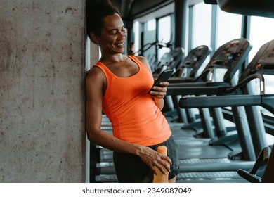 Woman standing in the gym and holding her smartphone in one hand and bottle of water in another. She is texting to someone or reading something cheerfully. - Powered by Shutterstock