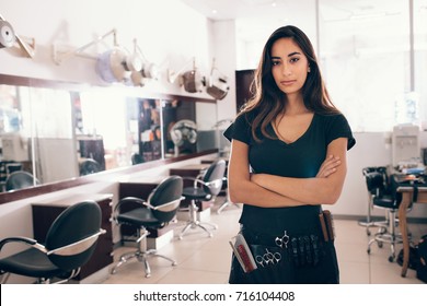 Woman standing with hands folded in her salon. Professional hairdresser with all the hairdressing accessories tied to her waist.