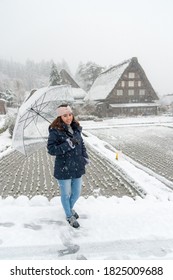 Woman Standing And Holding Transparent Umbrella In The Winter And Snow Is Falling. With Background Of Ancient House At Shirakawa-go Village At Gifu, Japan. With Copy Space