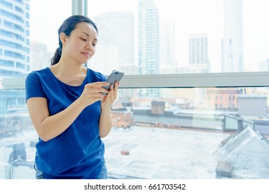 Woman standing indoors by window, day, urban scene, using mobile phone. asian, long hair, adult