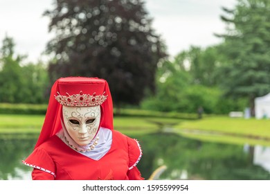 A woman standing in mask and red masquerade costume agains the pond background