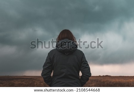 Woman standing in meadow, looking at the horizon and dark dramatic stormy clouds, rear view selective focus