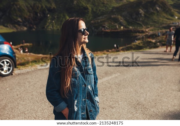 Woman standing in the middle of the\
road looking at the mountain on which she is going to climb\
together with the group of other tourists. Summer road\
trip.