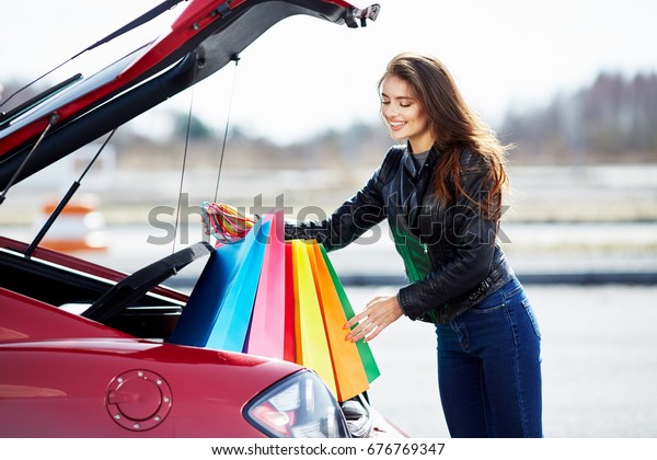 Woman standing near the red car, happy\
driver. Woman looking down and smiling, outside the car. Happy\
woman putting colorful bags after shopping into\
car
