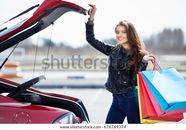 Woman standing near the red car, happy driver.\
Girl looking at camera and smiling, outside. Happy woman holding\
colorful bags after\
shopping
