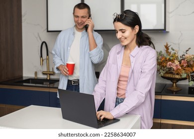 A woman is standing near a table with a laptop, in the background a man is talking on the phone - Shutterstock ID 2258634007