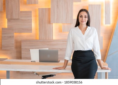 Woman Standing Near Table In Wooden Office Interior Room, Wearing White Shirt And Black Skirt. Female Secretary Looking At The Camera, Smiling. Concept Of Office Worker