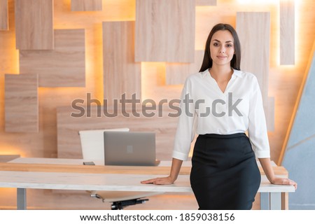 Woman standing near table in wooden office interior room, wearing white shirt and black skirt. Female secretary looking at the camera, smiling. Concept of office worker